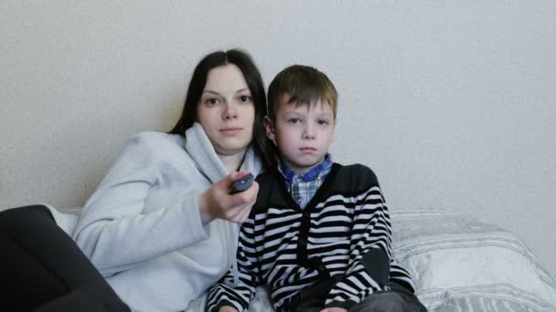 Viendo la tele tirada en el sofá. Mamá e hijo miran televisión juntos y sonríen. Mamá cambia de canal . — Vídeos de Stock