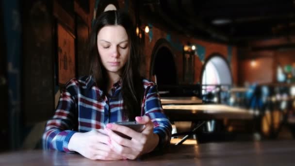 動揺して女性がメッセージを送信またはカフェに座って携帯電話でインターネットを使用します。格子縞のシャツを着た. — ストック動画