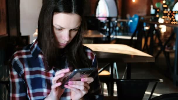 Mujer molesta envía un mensaje o uso de Internet en el teléfono sentado en un café. Vestido con una camisa a cuadros . — Vídeo de stock