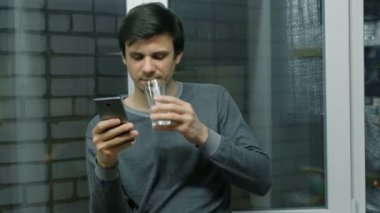 Adam üstünde hareket eden pencere balkon yakın duran ve bir su içme telefon internet sayfalarının gözatma.