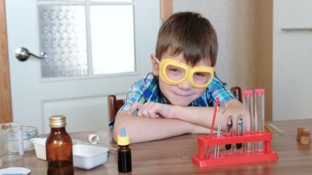 Expériences en chimie à la maison. Garçon en chemise à carreaux s'assoit à la table et va pratiquer des expériences de chimie. Souriant et regardant la caméra . — Video