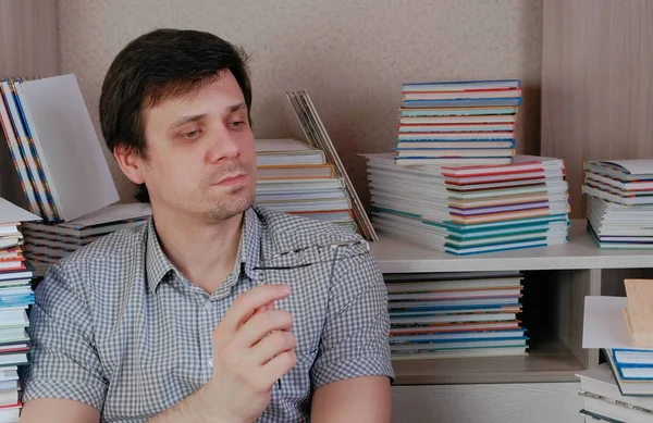 Joven hombre morena sentado entre los libros girando las gafas en sus manos y piensa . — Foto de Stock