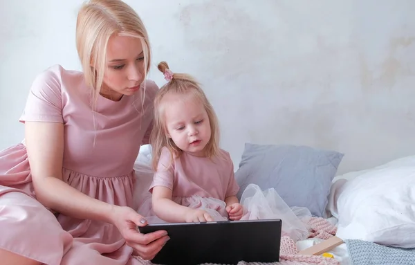 Mamma och dotter titta i tablett. Ung attraktiv blond kvinna med hennes lilla charmiga dotter i rosa klänningar du tittar på något i tablett. — Stockfoto
