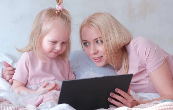 Young attractive blond woman teaches her little charming daughter in pink dresses using a tablet laying on bed.