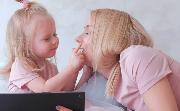 Ung attraktiv blond kvinna lär sin lite charmiga dotter i rosa klänningar använda en surfplatta som lägger på säng. Närbild porträtt. — Stockfoto