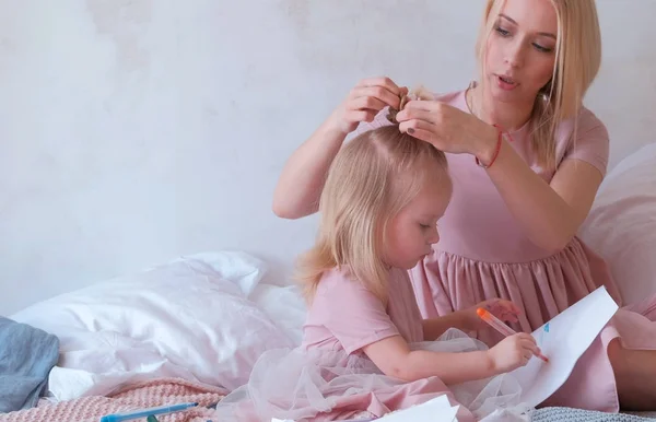 Ung attraktiv blond kvinna flätar hår till sin lilla charmiga dotter i rosa klänningar medan hon målar med filt-pen. — Stockfoto