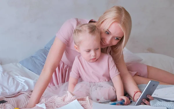 Ung attraktiv blond mamma visar något i mobiltelefon till sin lilla charmiga dotter i rosa klänningar. Eller gör Tuesuti — Stockfoto