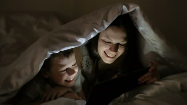 若い女性のお母さんとタブレット、毛布の下で一緒に面白い映画を見て彼女の息子. — ストック動画