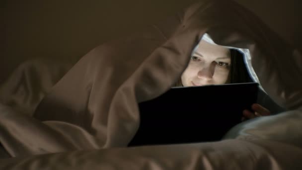Ung kvinde ser en sjov film på tablet under tæppet . – Stock-video