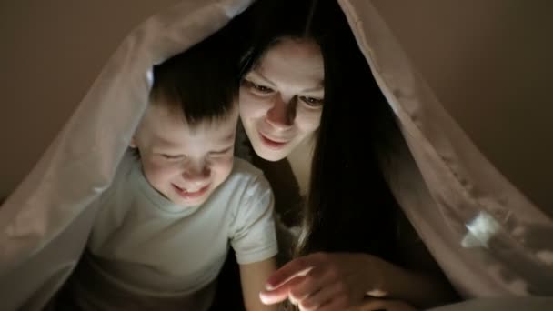 Mujer joven mamá y su hijo viendo una película divertida juntos en la tableta y reír bajo la manta . — Vídeos de Stock