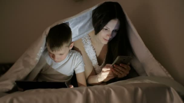 Young beautiful woman mom browsing internet in her phone while her son watching cartoon on tablet. — Stock Video