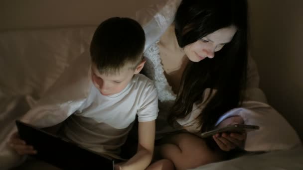 Young beautiful woman mom browsing internet in her phone while her son watching cartoon on tablet. — Stock Video