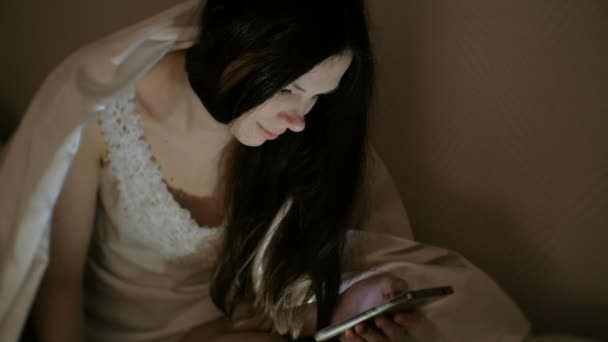 Young beautiful woman brunette browsing internet in her mobile phone in bed before sleeping. — Stock Video