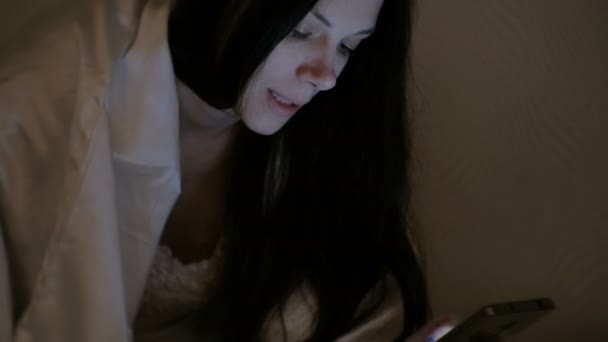 Giovane bella donna bruna parla tramite collegamento video nel suo telefono cellulare a letto prima di dormire e sorride . — Video Stock