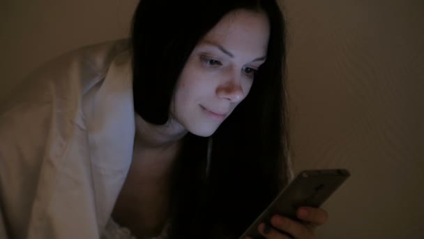 Mooie jongedame brunette spreekt door videoverbinding in haar mobiele telefoon in bed voordat slapen en glimlacht. — Stockvideo