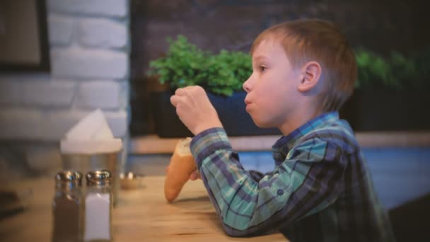 Хлопчик їсть багет і дивиться телевізор у кафе. Вид збоку . — стокове відео