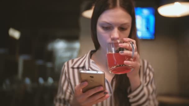 Young beautiful brunette woman watch something in mobile phone, smiles and drinks red herbal tea in cafe. — Stock Video
