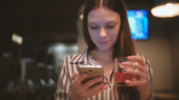 Jeune belle femme brune regarder quelque chose dans le téléphone mobile, sourit et boit tisane rouge dans le café . — Video