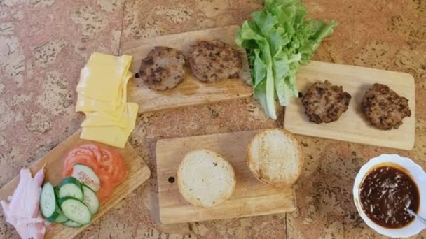 Products for preparation of burgers: buns, tomatoes, cucumbers, cutlets, cheese, salad, sauce, bacon on the table. Top view. — Stock Video