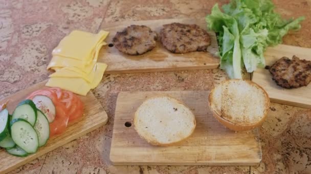 Prodotti per preparazione di hamburger: focacce, pomodori, cetrioli, costolette, formaggio, insalata, salsa, pancetta sul tavolo . — Video Stock
