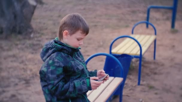 Menino joga um jogo em seu telefone celular sentado no parque em um banco . — Vídeo de Stock