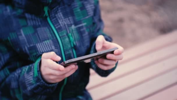 Jongen speelt een spel op zijn mobiele telefoon in het Park op een bankje zitten. Close-up jongens handen. — Stockvideo