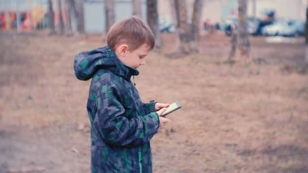 Pojke ringer upp ett nummer och talar på en mobiltelefon i parken. — Stockvideo