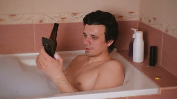 Ung man tar ett bad med skum, surfar internet på mobiltelefon, dricker öl från flaska. — Stockvideo