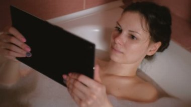 Genç güzel esmer kadın banyo ve saatler tablete video alır. Yakın çekim yüz.