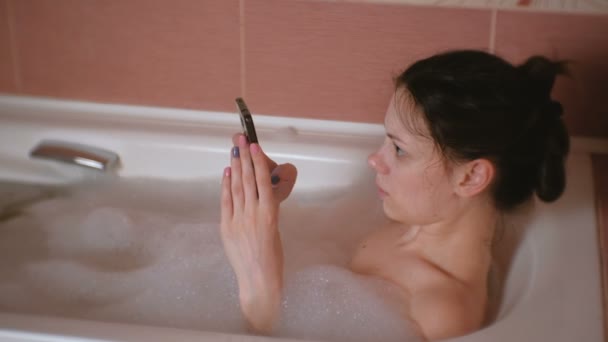 Молода красива брюнетка приймає ванну, переглядаючи Інтернет у мобільному телефоні . — стокове відео