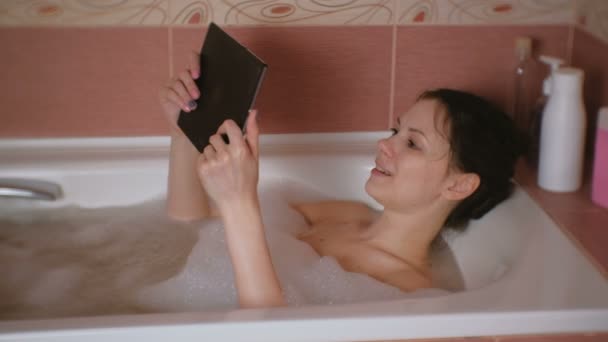 Jovem bela mulher morena toma banho e assiste vídeo no tablet . — Vídeo de Stock