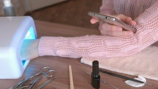Женщина сушит ногти Шеллаком в ультрафиолетовой лампе и просматривает интернет в мобильном телефоне. Рука крупного плана . — стоковое видео