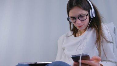 Kadın gözlük ve kulaklık smartphone ve koltukta oturan tablet kullanarak çevrimiçi mağazada alışveriş yapar.