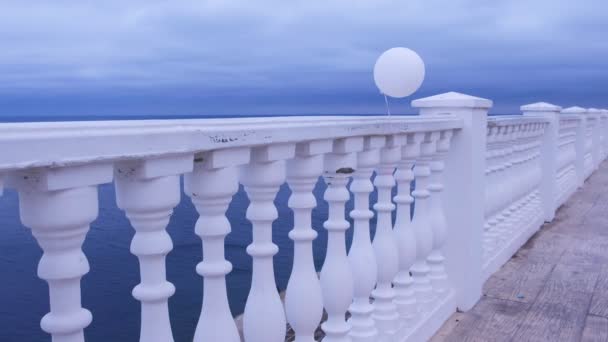 Bílý balon ve větru přivázaný k plotu na mořském nábřeží za oblačného dne. — Stock video