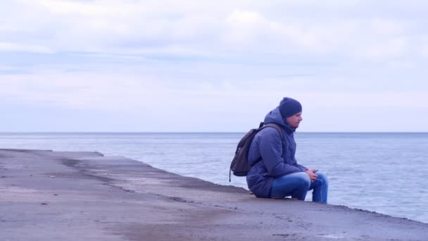 Man tourist sits on old sea waterfront and looks at sea in winter with phone. — Stock Video