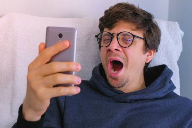 Gözlüklü uykulu adam esneiyor ve koltukta oturan akıllı telefonda sosyal medyada geziniyor.