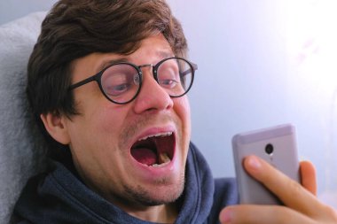 Gözlüklü adam dişlerini cep telefonunda kamera inceliyor.