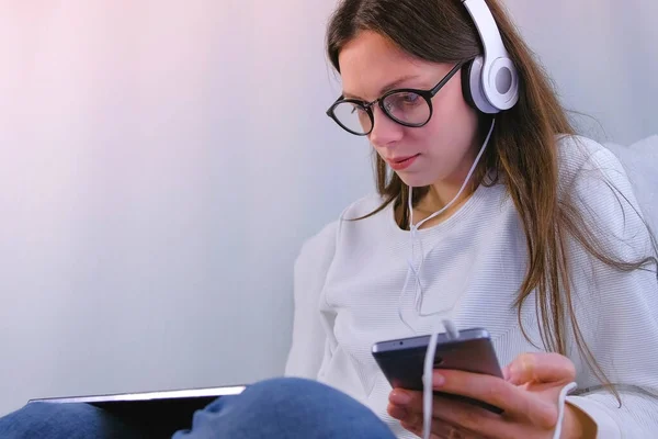 Woman in glasses and headphones makes purchases in the online store using smartphone and tablet sitting in armchair. — Stock Photo, Image