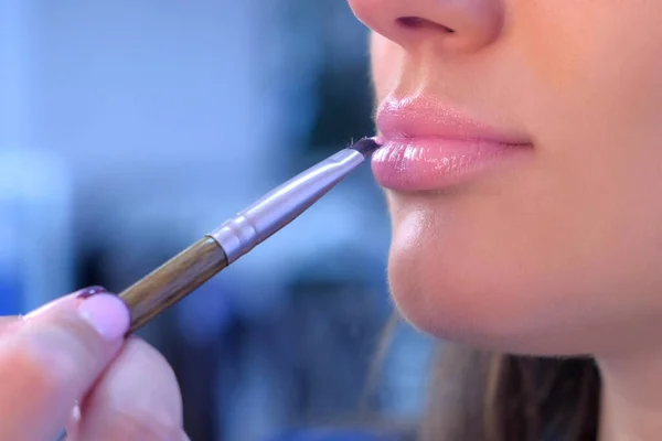 Makeup artist applies lip gloss uses brush on girl model lips makes make up.