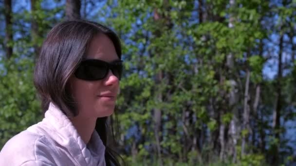 Портрет женщины откладывает солнцезащитные очки, сидя в лесу на берегу реки . — стоковое видео