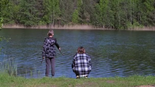春の暖かい日に森の近くの川岸で休んでいる母と息子、バックビュー. — ストック動画