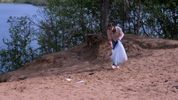 Dobrovolnice čistí písečnou pláž v blízkosti jezera, sbírá odpadky v tašce. — Stock video