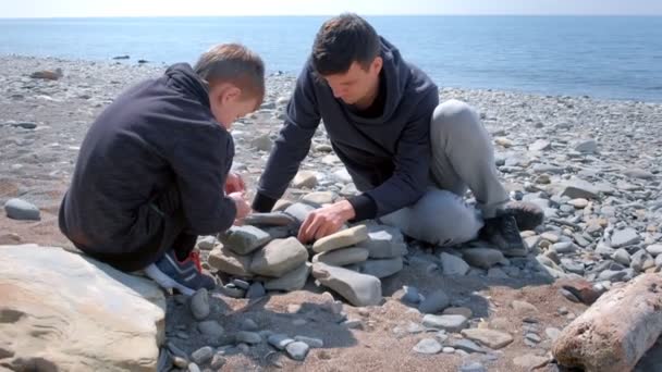 Baba ve oğlu deniz taşı sahilde ahşap sopalarla taş brazier üzerinde şenlik ateşinde kömürleri tutuşturmak vardır. — Stok video