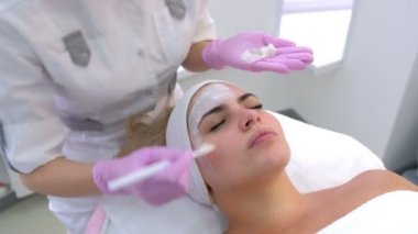 Kozmetik uzmanı, güzellik kliniğinde kadın müşteri yüzüne beyaz maske takıyor..