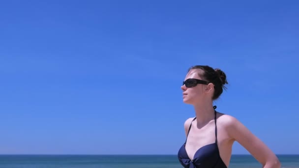 肖像画女性日光浴は海と空の背景に水着でビーチに立つ. — ストック動画