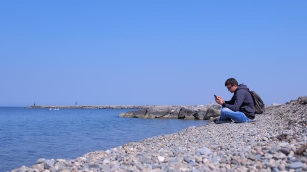 Adam gezgin deniz taş plajda oturan ve akıllı telefon tarama, yan görünüm. — Stok video