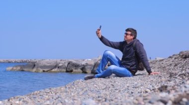 Adam gezgin deniz taş plajda oturur ve tatil selfie akıllı telefon yapar.