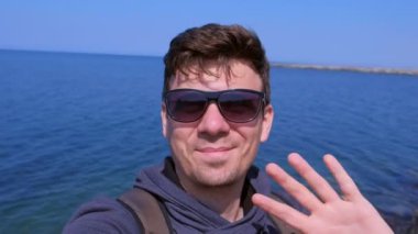Adam görüntülü sohbetler selfie smartphone deniz plajı dalgalar el selamlar ve gülümsüyor.