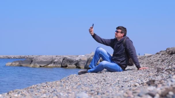남자 여행자는 바다 돌 해변에 앉아 휴가에 셀카 스마트 폰을 만든다. — 비디오