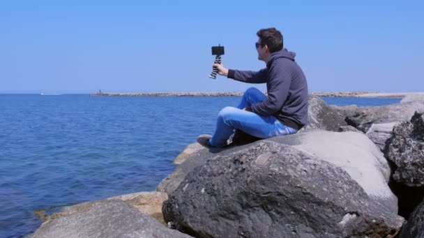 남자 회담 화상 채팅 에 스마트 폰 앉아 에 거대한 돌 에 바다 해변 에 휴가. — 비디오
